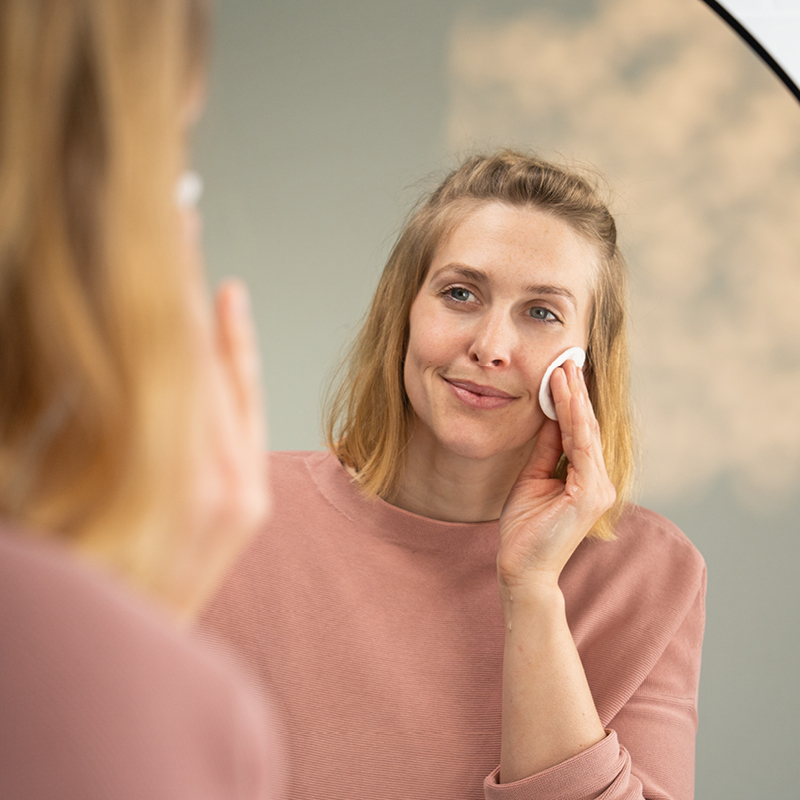 Frau trägt lächelnd vor einem Spiegel ein Produkt mit einem Wattepad auf ihr Gesicht auf