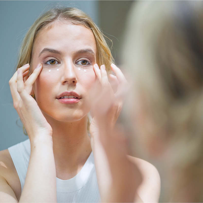 Frau schaut sich in einem Spiegel an und klopft ihre Augenpflege ein.