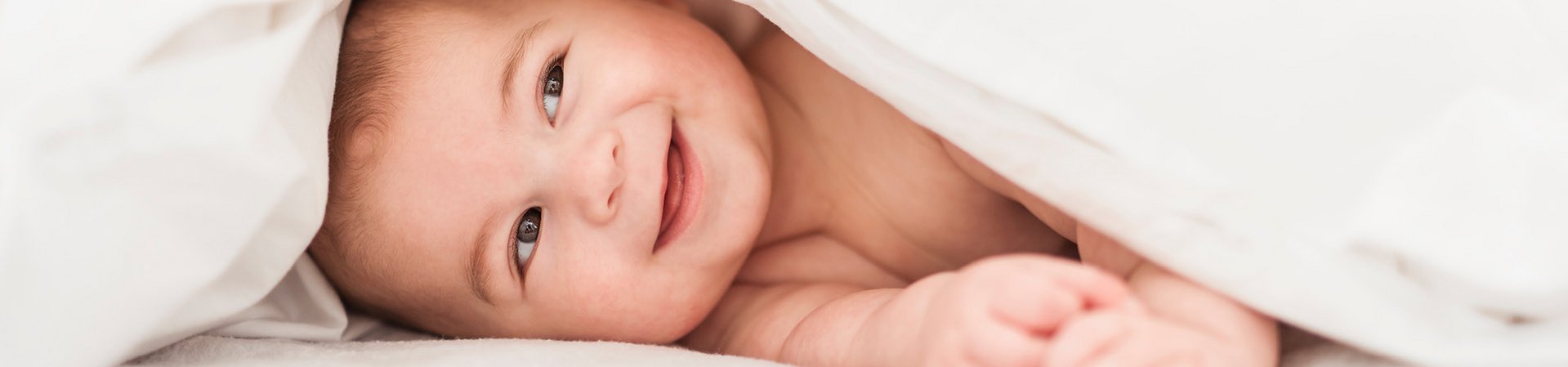 Babyhaut bedarf besonders verträglicher Hautpflege Lächelndes Baby unter einer weißen Decke