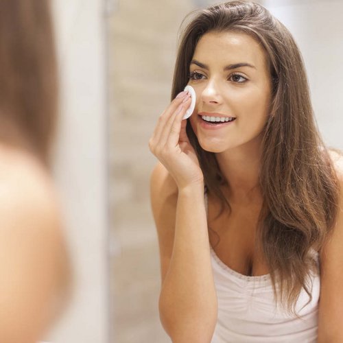 Eine junge Frau fährt sich mit einem Wattepad über das Gesicht und schaut dabei in den Spiegel.