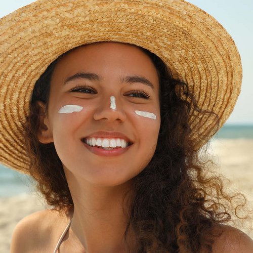 Eine junge brünette Frau mit Sonnencreme im Gesicht trägt einen Strohhut am Strand.